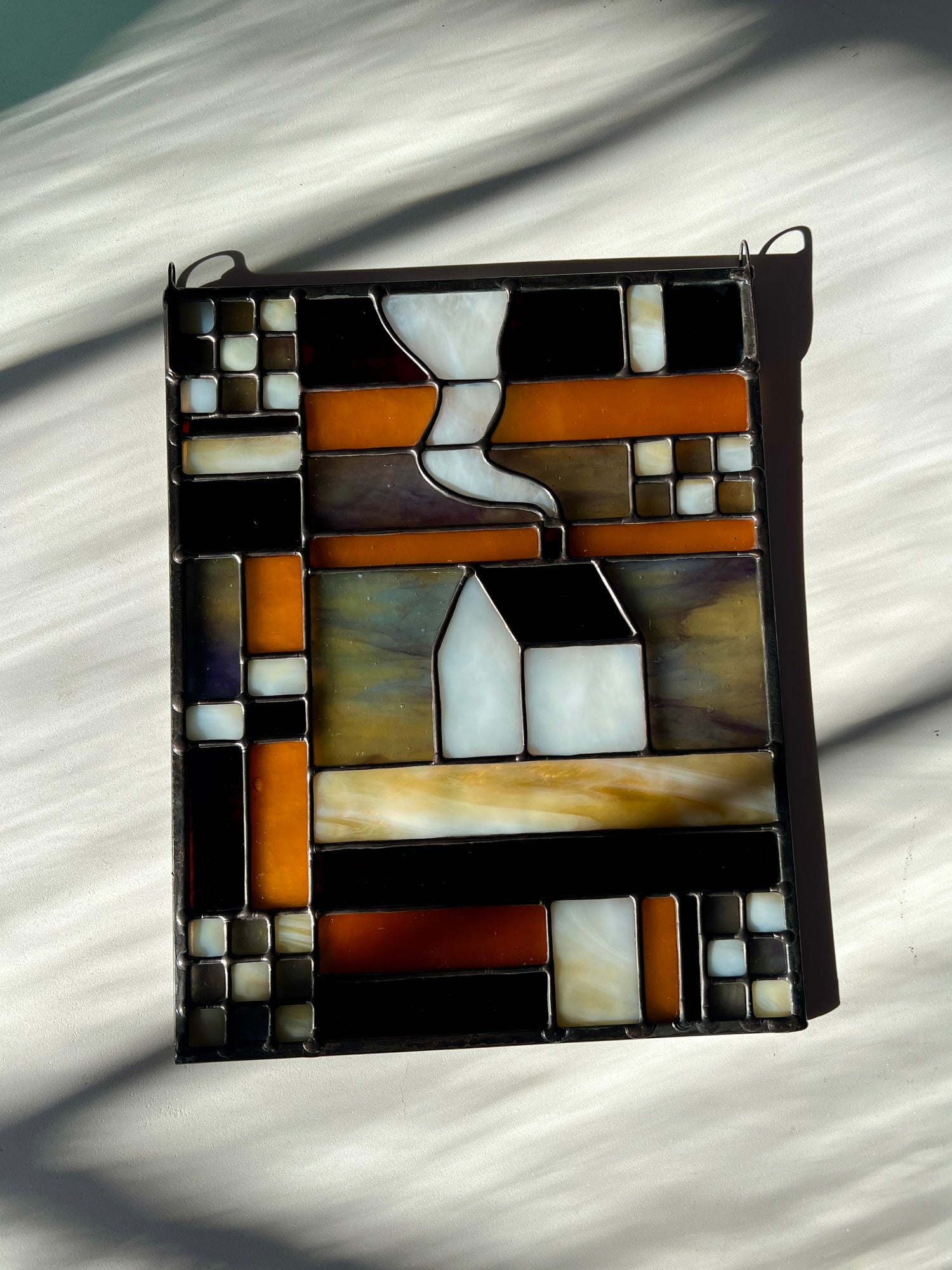 Stained glass textile inspired panel depicting a home in the center in shades of purple, white, brown, and amber, resting on a white surface with sunlight and shadows cast across