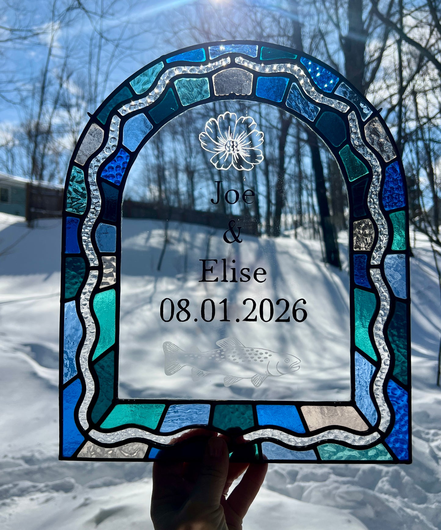 Stained glass panel in the shape of an arch with a multicolor blue, teal, turquoise, and pink border. Within the border is squiggle detail in clear textured glass. The center of the arch is clear uncolored glass, with personalized text and hand etched details of a cosmos flower and trout. The piece is held by a hand outside with bright sunlight providing backlighting.