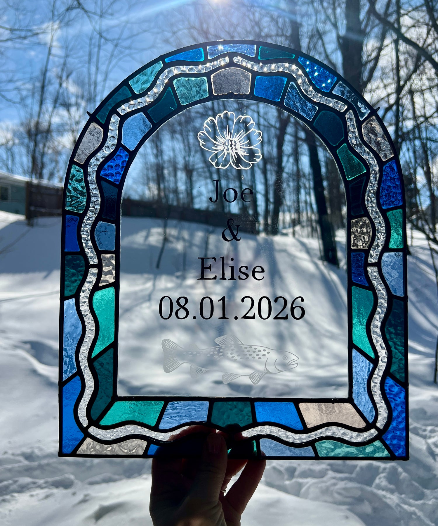 Stained glass panel in the shape of an arch with a multicolor blue, teal, turquoise, and pink border. Within the border is squiggle detail in clear textured glass. The center of the arch is clear uncolored glass, with personalized text and hand etched details of a cosmos flower and trout. The piece is held by a hand outside with bright sunlight providing backlighting.