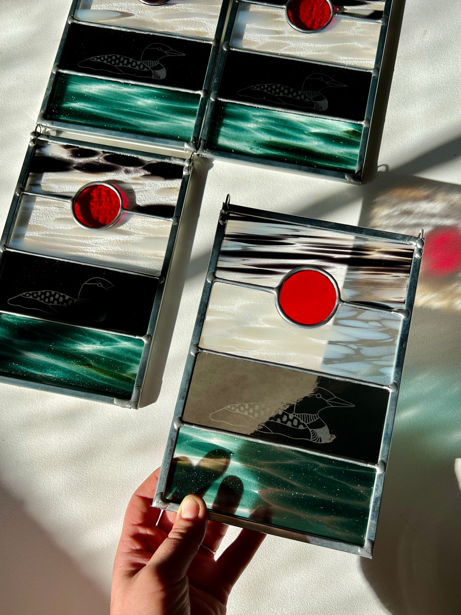 a hand holding a stained glass panel with black, white, clear, teal, and red glass with a loon etched on the surface, with three similar panels resting on a white surface in the background
