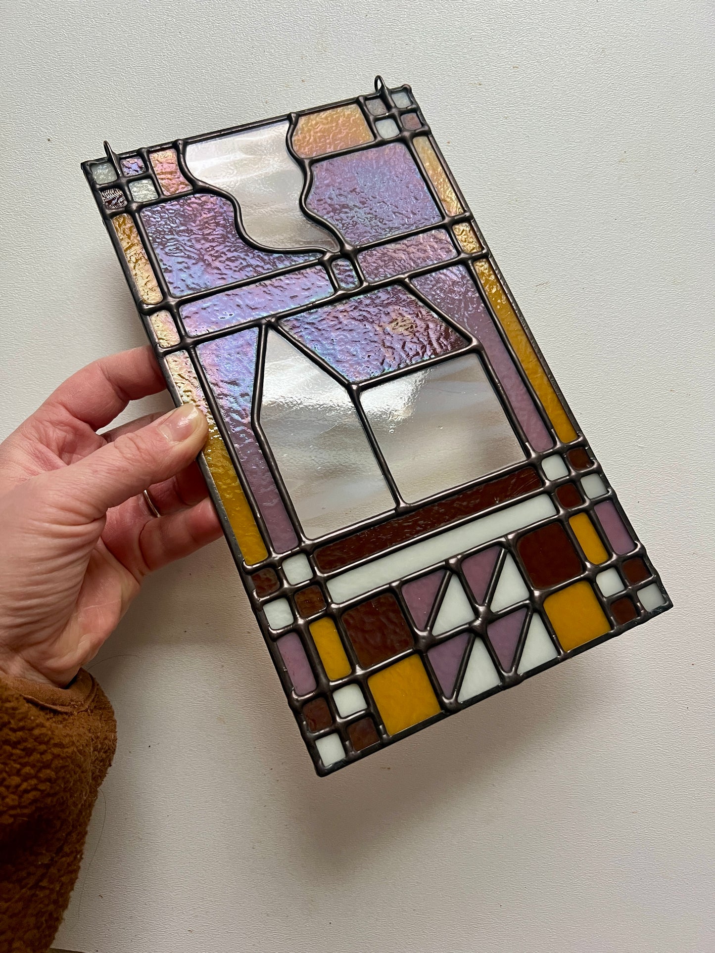 Stained glass textile inspired panel depicting a home in the center in shades of purple, white, brown, and bronze, held by a hand at an angle against a white surface, highlight slight 'oil slick' effect of the glass