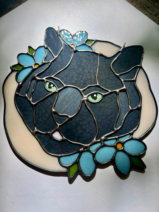 A stained glass art piece depicting a black cat's face with green eyes, surrounded by light blue flowers and a cream-colored free form background, set against a plain white background.