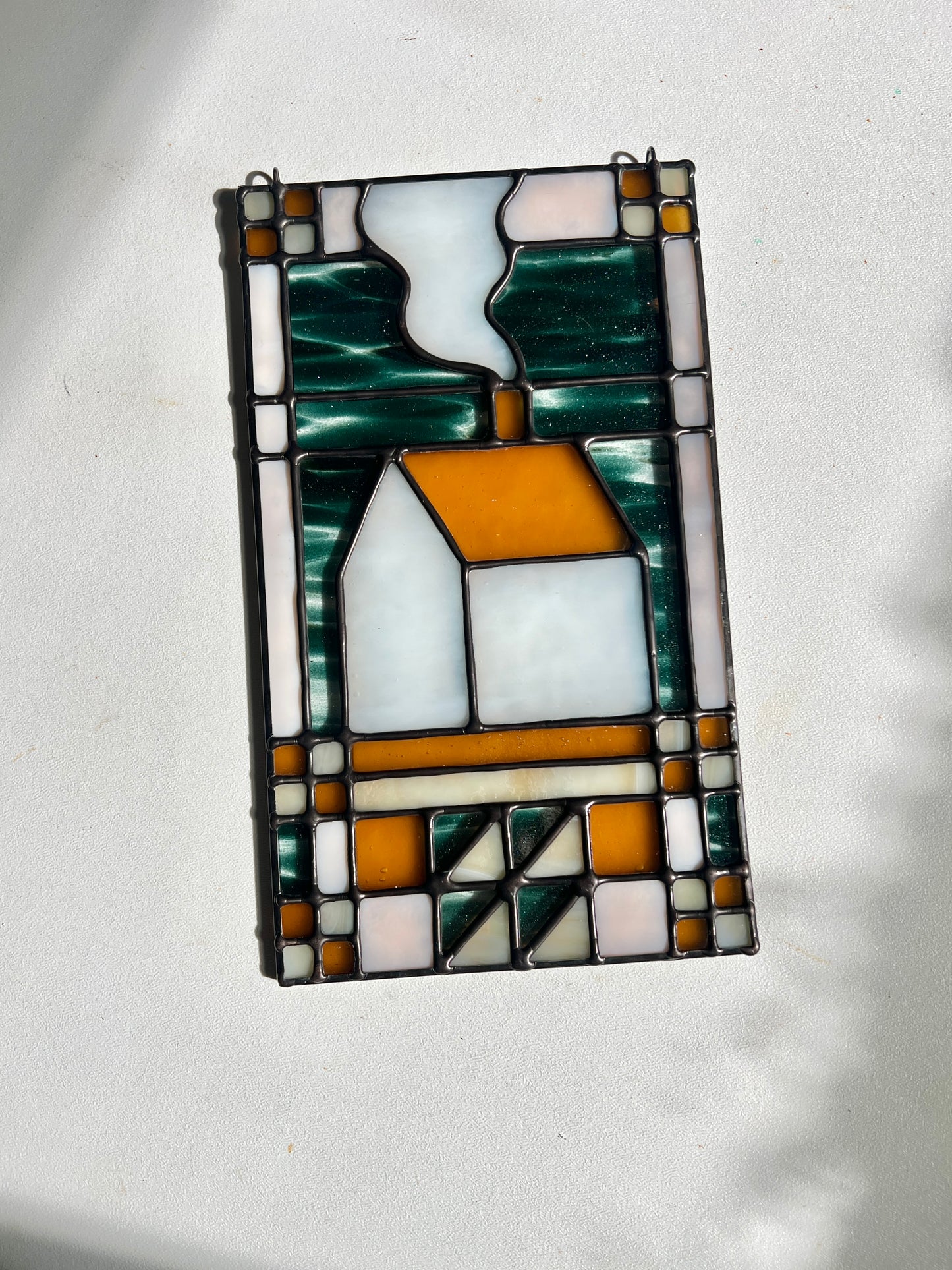 Stained glass textile inspired panel depicting a home in the center in shades of teal, amber, white, and beige, resting against a white surface in bright sunlight