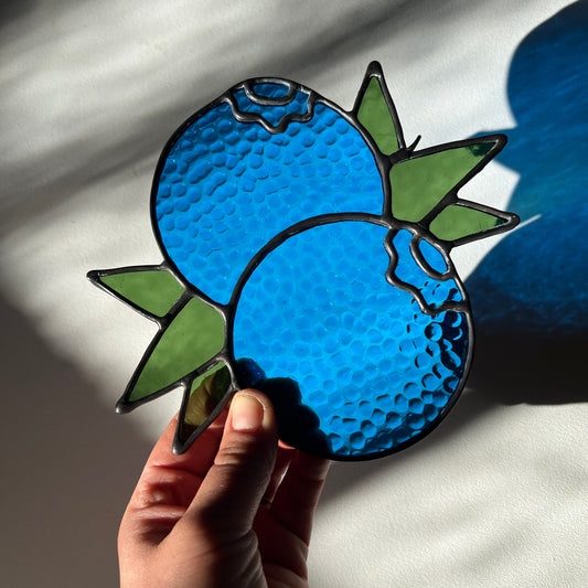 hand holding a textured stained glass suncatcher depicting a small bunch of blueberries with leaves