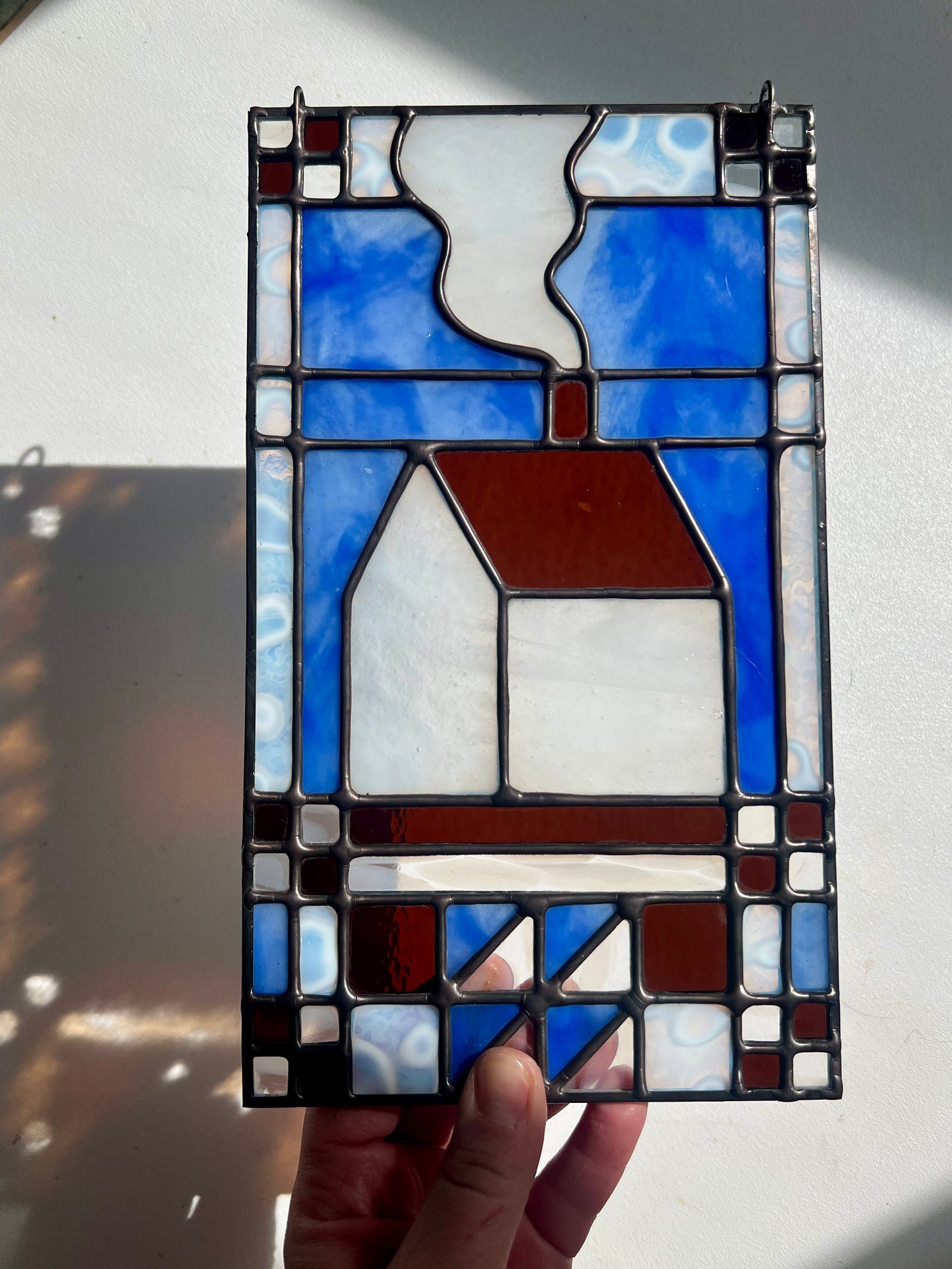 Stained glass textile inspired panel depicting a home in the center in shades of blue, brown, and white, held by a hand in front of a white surface with bright sunlight cast across