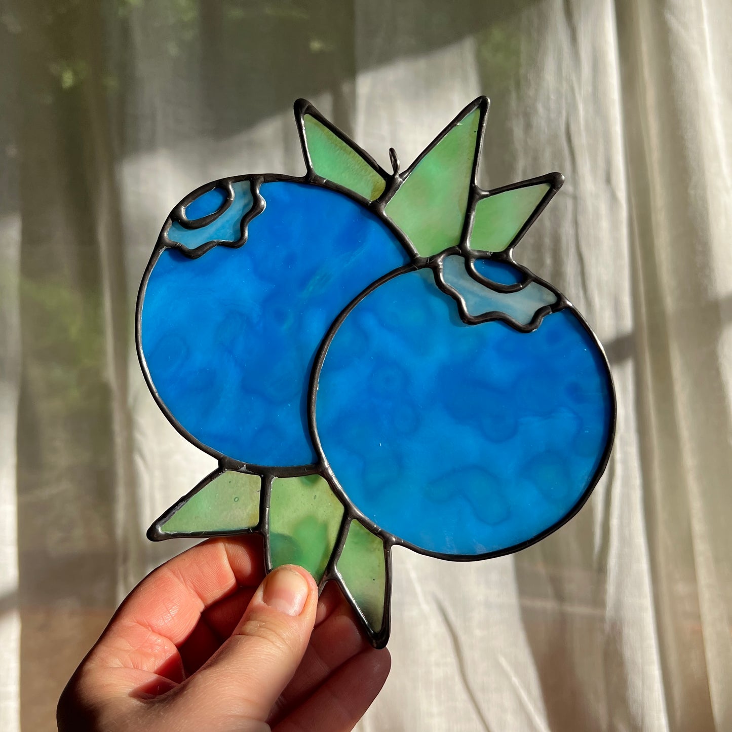 hand holding a mottled blue stained glass suncatcher depicting a small bunch of blueberries with leaves in front of a window