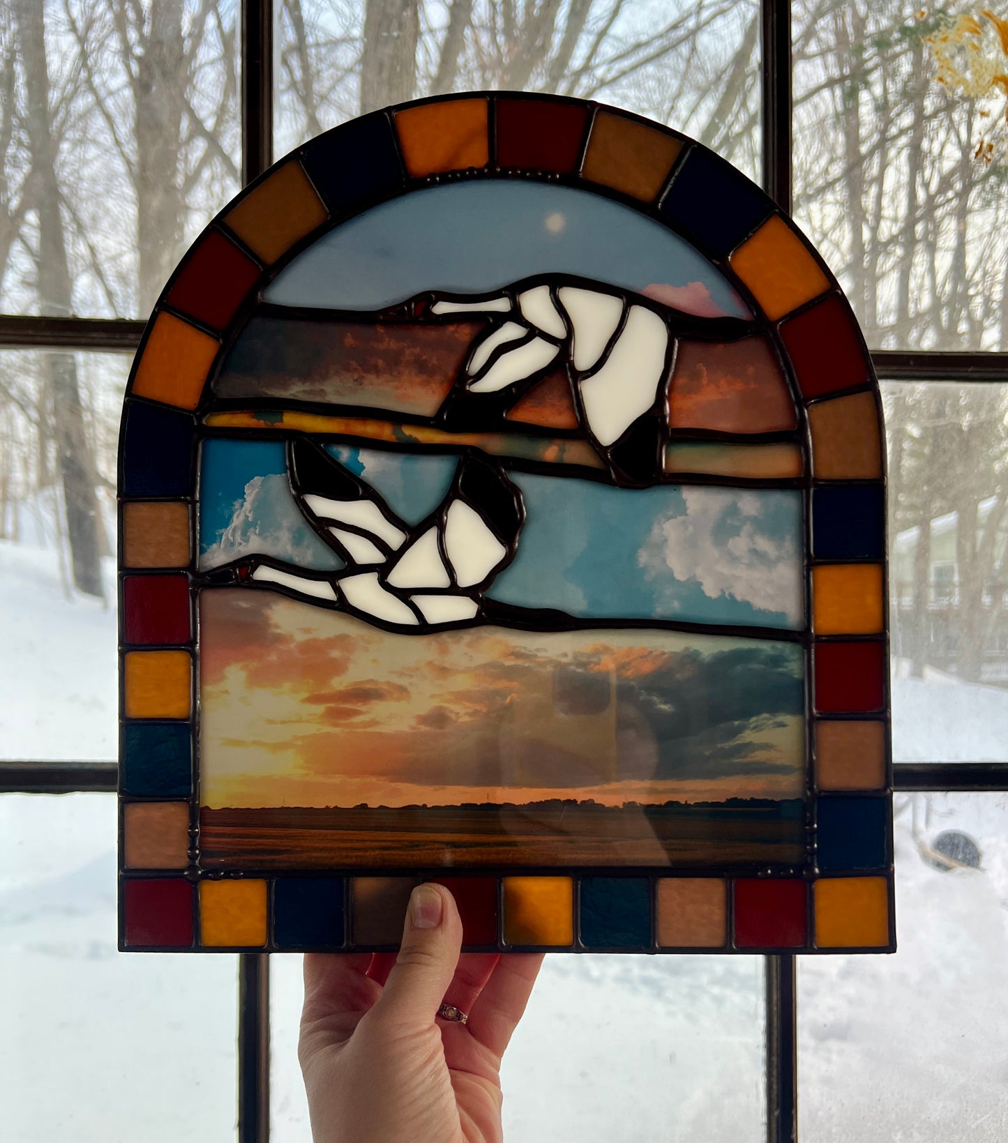 Stained glass panel with a landscape design created by collaged photographs and two cranes flying, held against a snowy window background