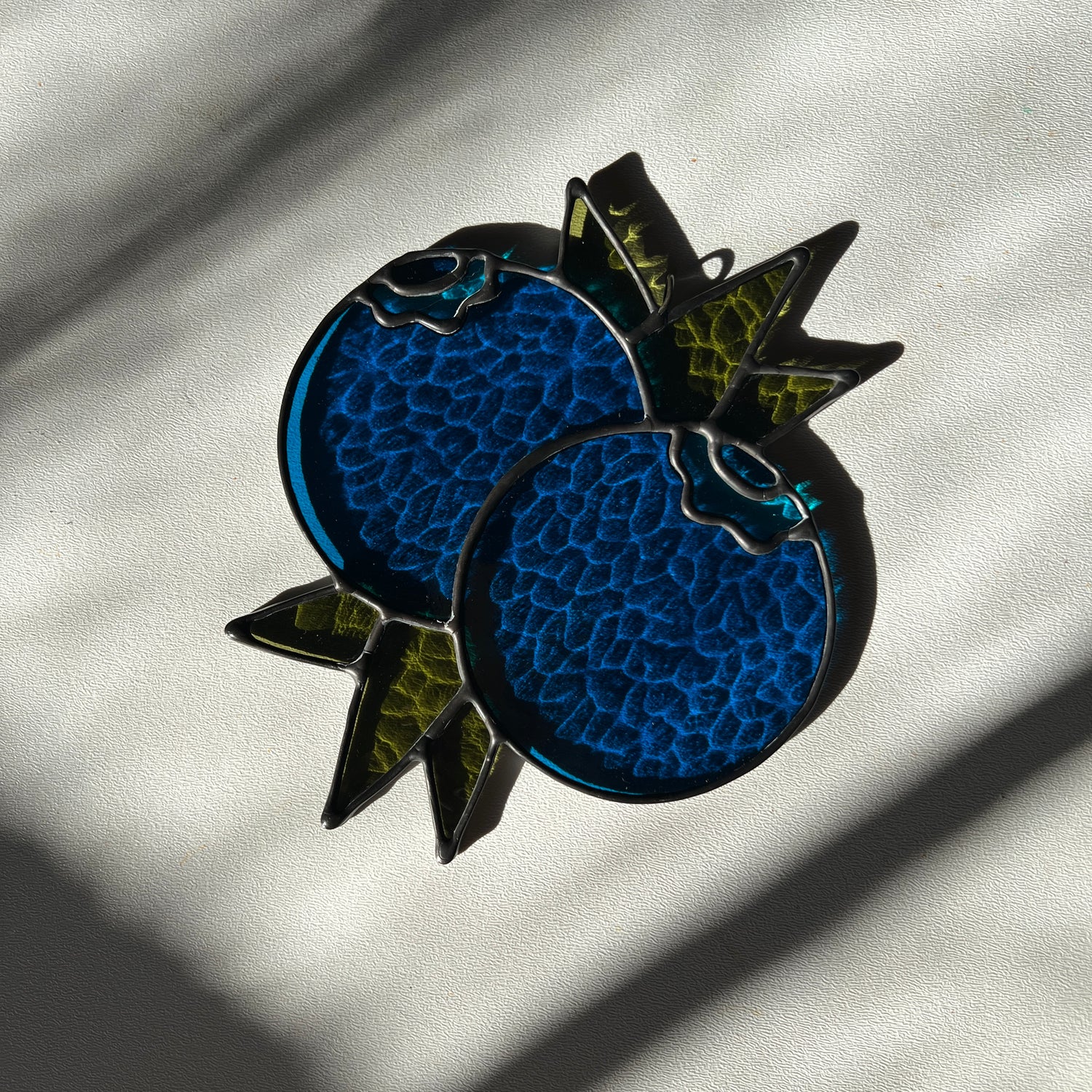 a textured blue stained glass suncatcher depicting a small bunch of blueberries with leaves resting on a white surface