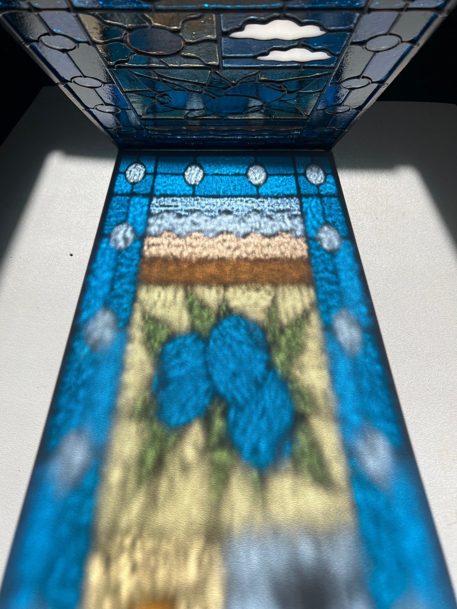 close up of a colorful refraction of a stained glass panel with a simple compartment style design featuring clouds, sun, water, and a small bunch of blueberries in the middle with a blue border