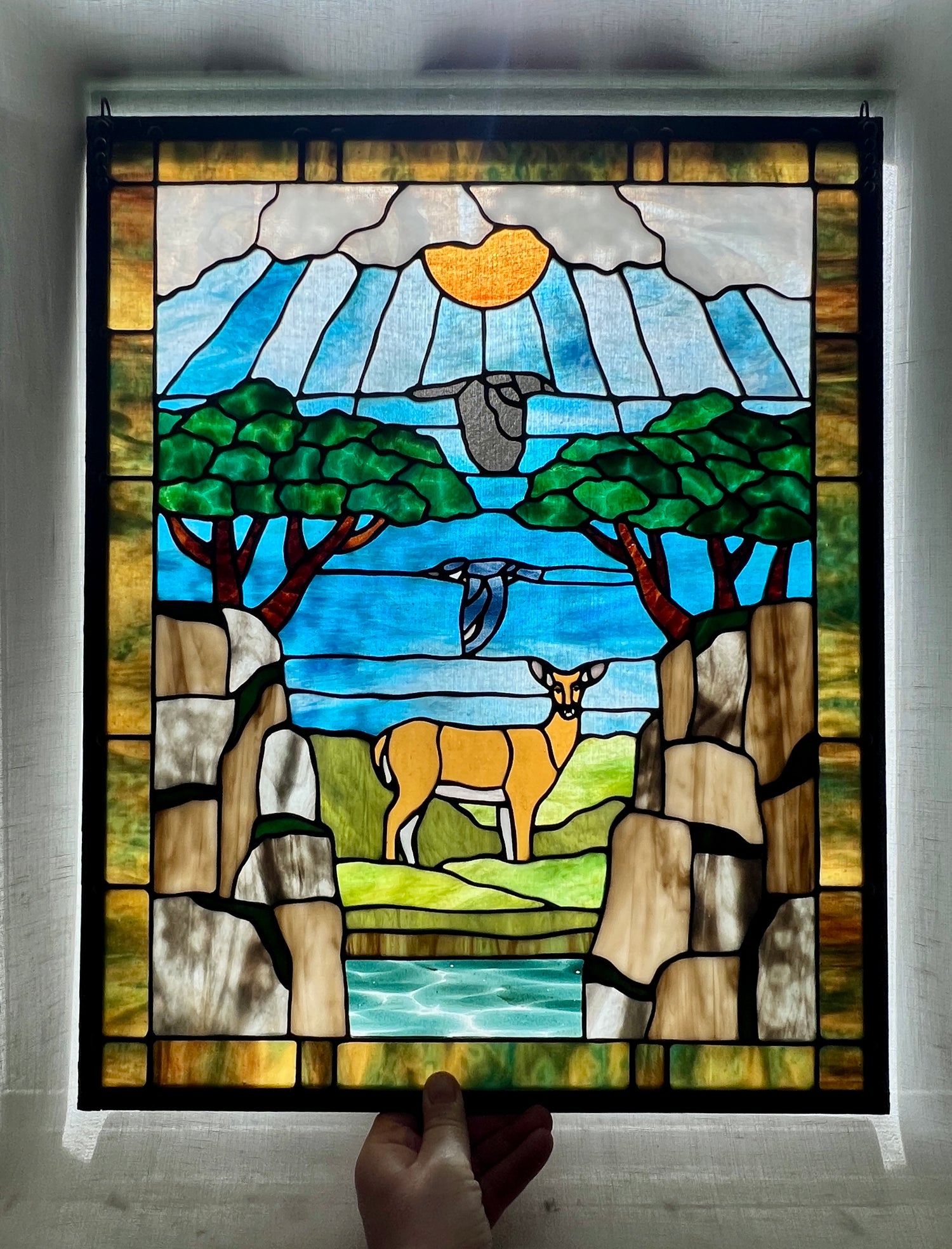 large stained glass panel depicting mossy, rocky cliffs and trees, with a deer, a kingfisher, and a raven standing amongst a river, greenery, and bright blue skies with some clouds and a bright yellow orange sun.