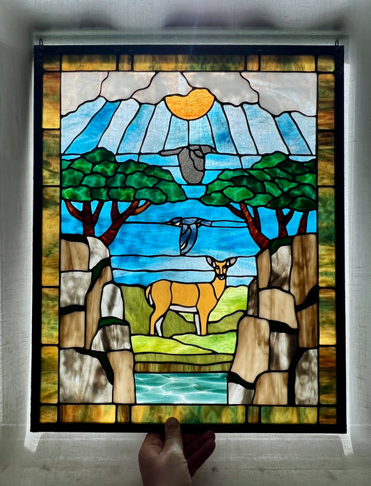 large stained glass panel depicting mossy, rocky cliffs and trees, with a deer, a kingfisher, and a raven standing amongst a river, greenery, and bright blue skies with some clouds and a bright yellow orange sun.