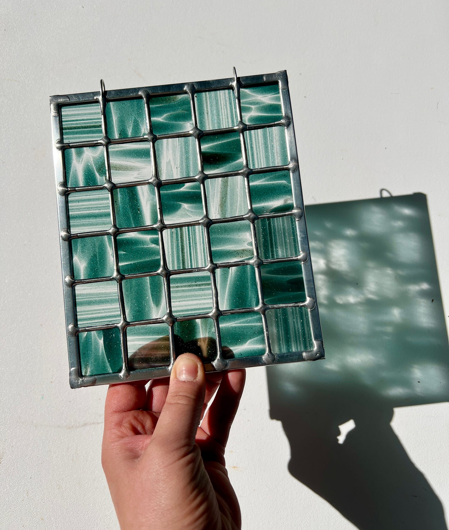 hand holding a stained glass suncatcher depicting streaky and mottled sparkly clear and teal glass arranged in a grid pattern, held above a white surface with sunlight casting a partial refraction
