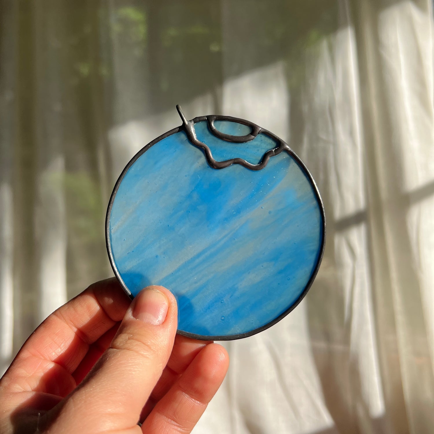 hand holding a light blue wispy stained glass blueberry in front of a window