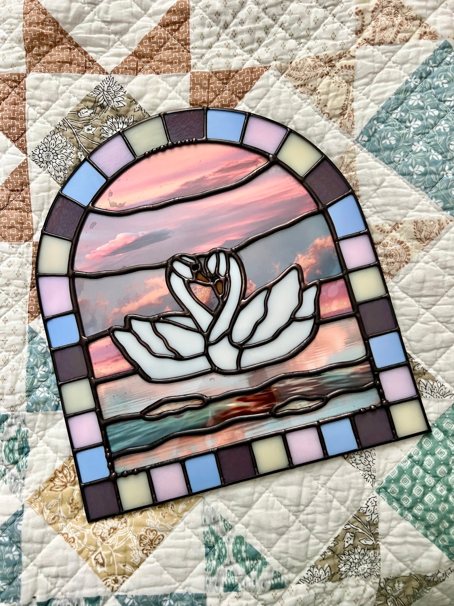 Stained glass panel with a colorful pastel arch border and two white swans in the center. Collaged photographs are used to create a sunset background. The panel is resting on top of a pastel quilt.