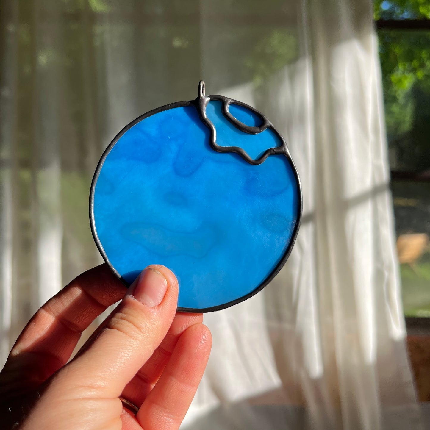 hand holding a light blue mottled stained glass blueberry in front of a window