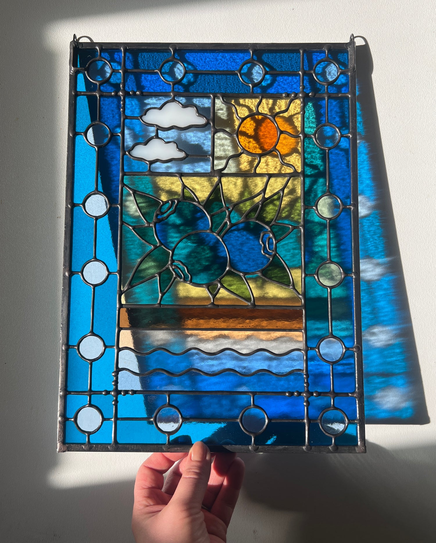 stained glass panel with a simple compartment style design featuring clouds, sun, water, and a small bunch of blueberries in the middle with a blue border held by a hand against a wall