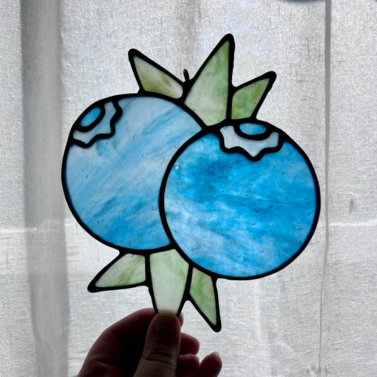 A hand holding a stained glass suncatcher depicting two blueberries and some leaves against a gently backlit white background.
