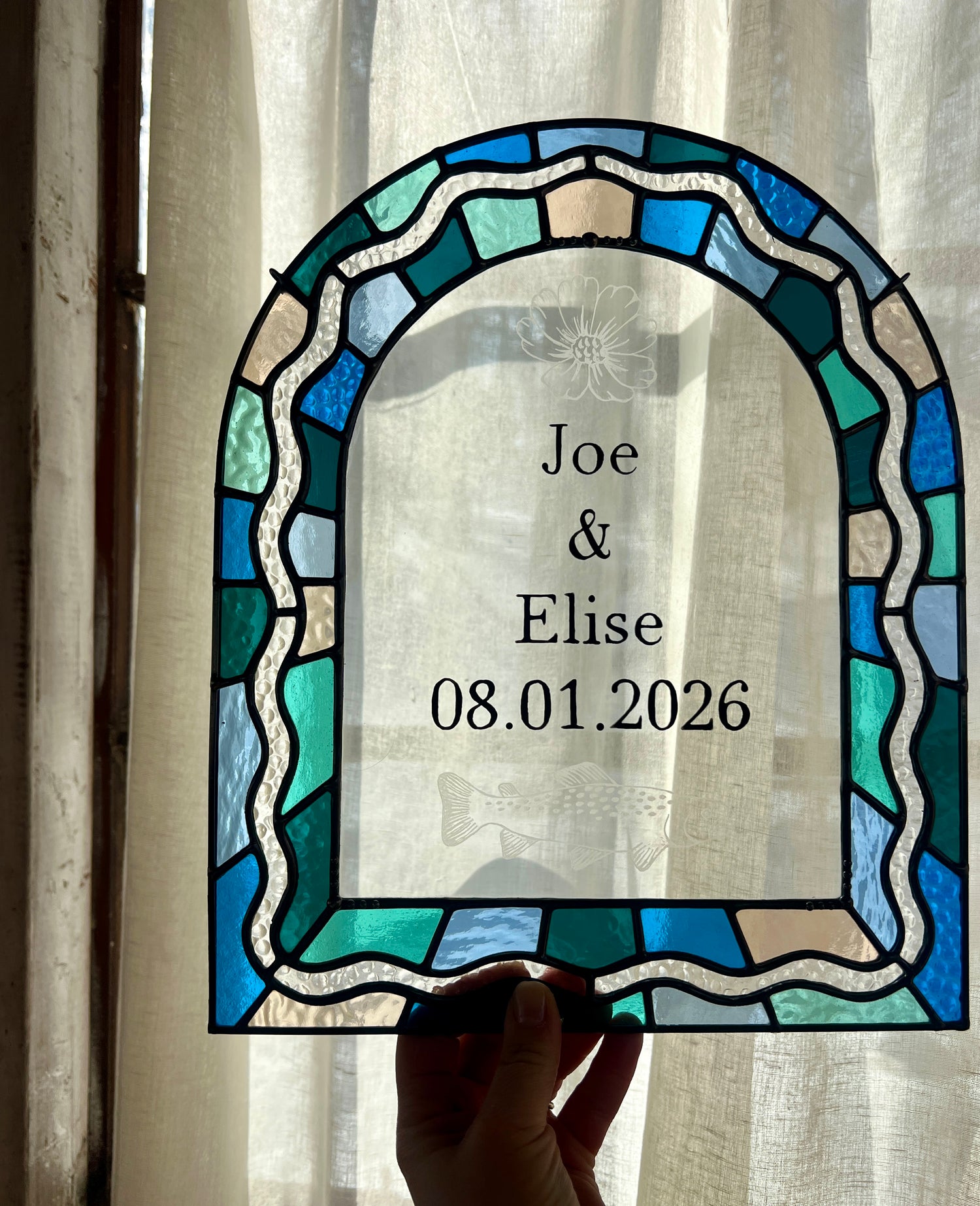Stained glass panel in the shape of an arch with a multicolor blue, teal, turquoise, and pink border. Within the border is squiggle detail in clear textured glass. The center of the arch is clear uncolored glass, with personalized text and hand etched details of a cosmos flower and trout. The piece is held by a hand against a sheer white current providing gentle backlighting.