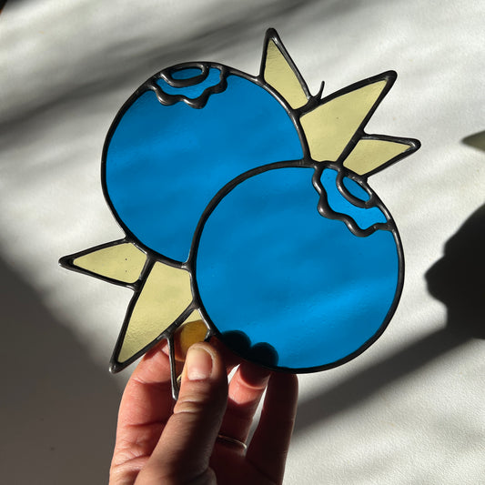 hand holding a stained glass suncatcher depicting a small bunch of blueberries with leaves
