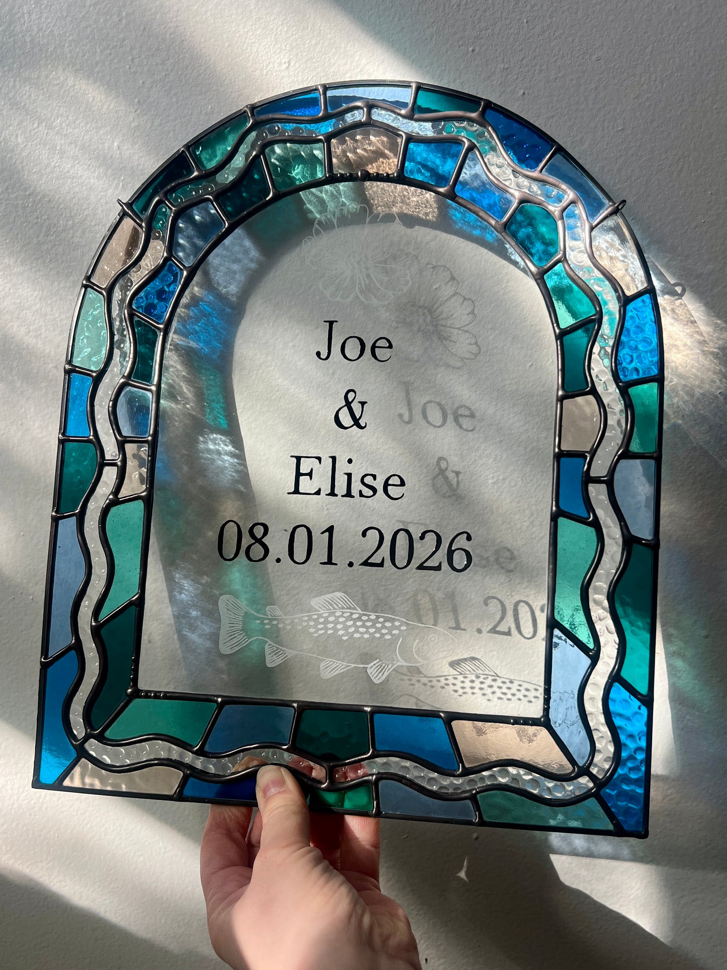 Stained glass panel in the shape of an arch with a multicolor blue, teal, turquoise, and pink border. Within the border is squiggle detail in clear textured glass. The center of the arch is clear uncolored glass, with personalized text and hand etched details of a cosmos flower and trout. The piece is held by a hand against a white wall, with gentle sunlight casting intermittent shadows and refractions.