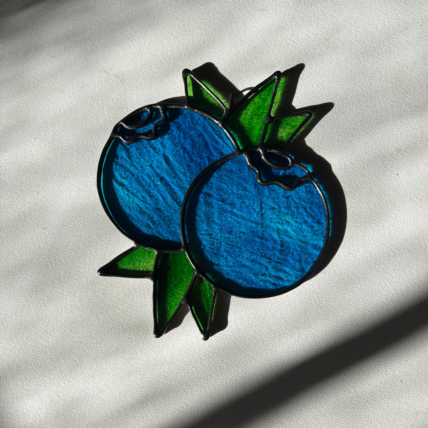  a wispy blue suncatcher depicting a small bunch of blueberries with leaves resting on a white surface