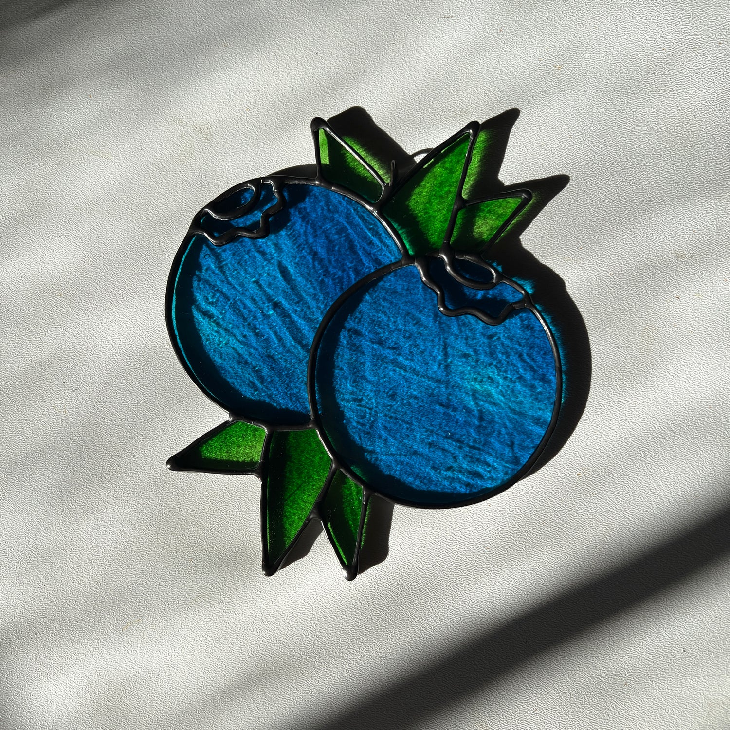  a wispy blue suncatcher depicting a small bunch of blueberries with leaves resting on a white surface