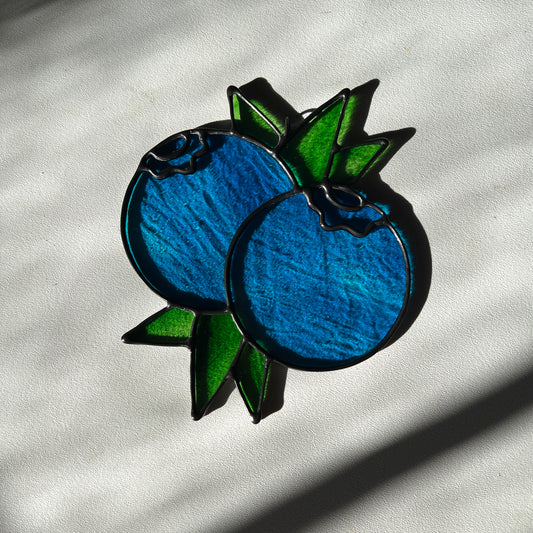  a wispy blue suncatcher depicting a small bunch of blueberries with leaves resting on a white surface