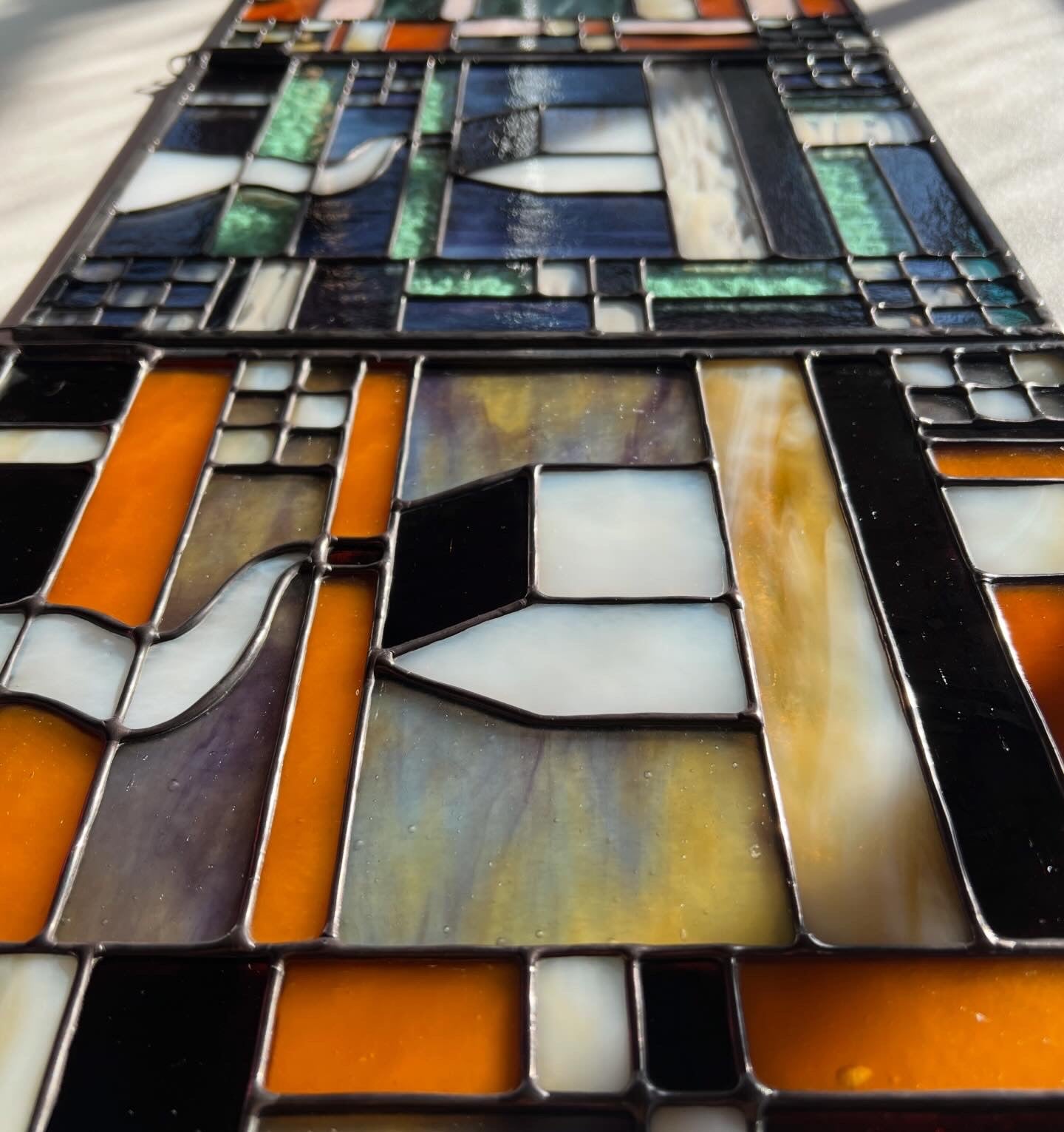 Stained glass textile inspired panel depicting a home in the center in shades of purple, white, brown, and amber, photographed at a close up angle to highlight smooth solder lines