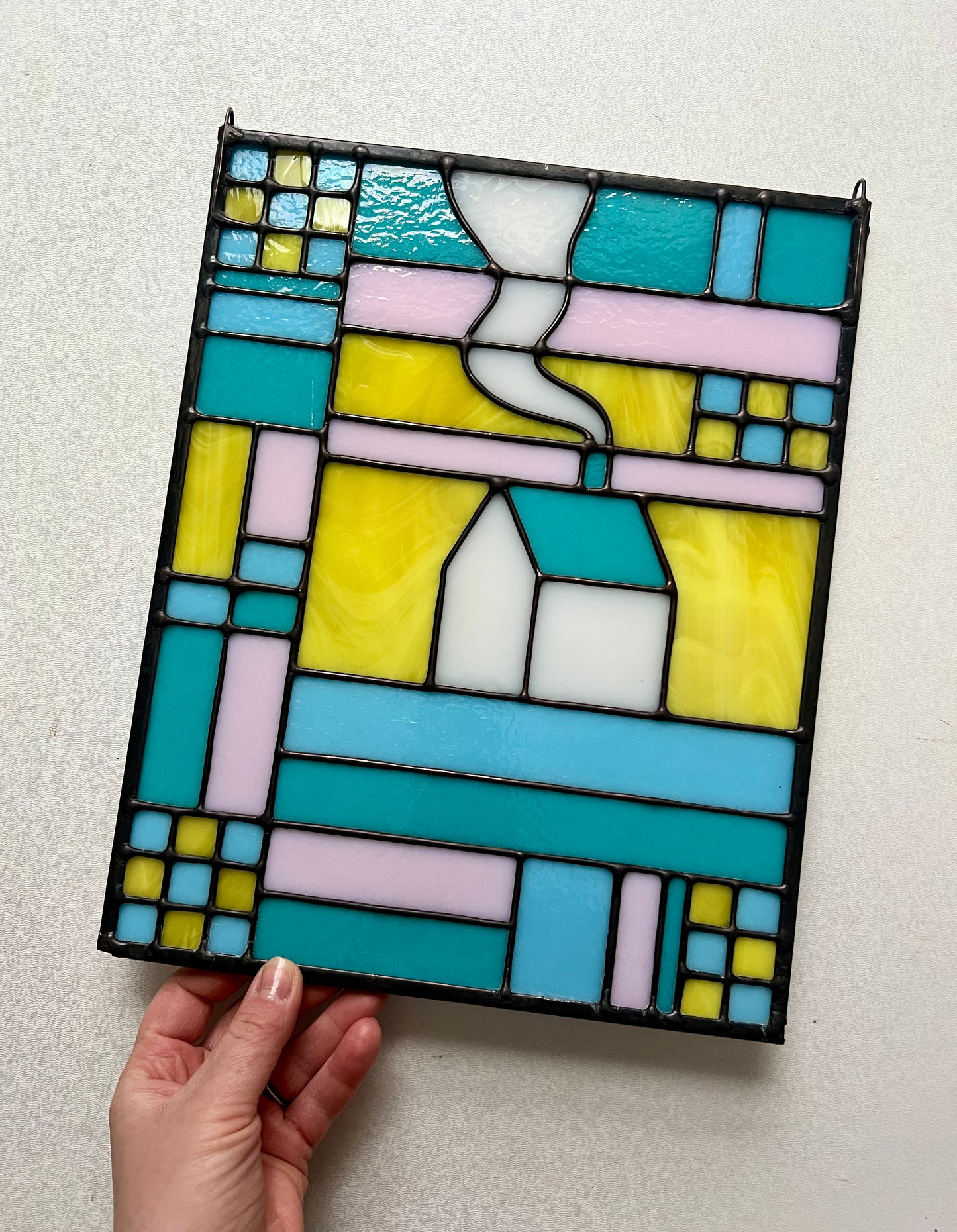 Stained glass textile inspired panel depicting a home in the center in shades of teal, pink, white, blue, and yellow, held by a hand against a white background