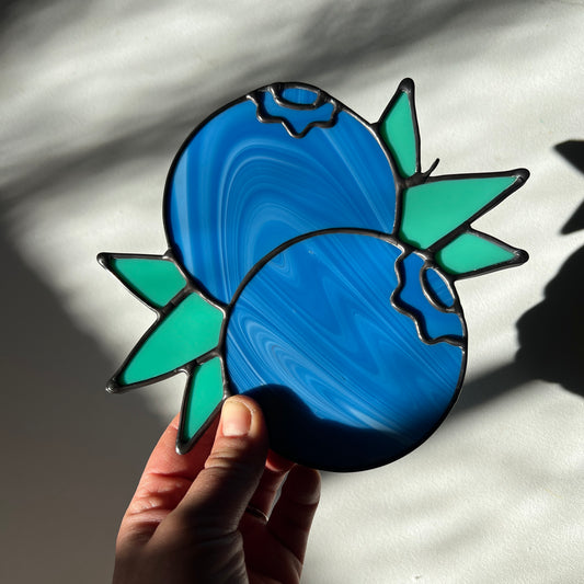 hand holding a streaky blue stained glass suncatcher depicting a small bunch of blueberries with leaves