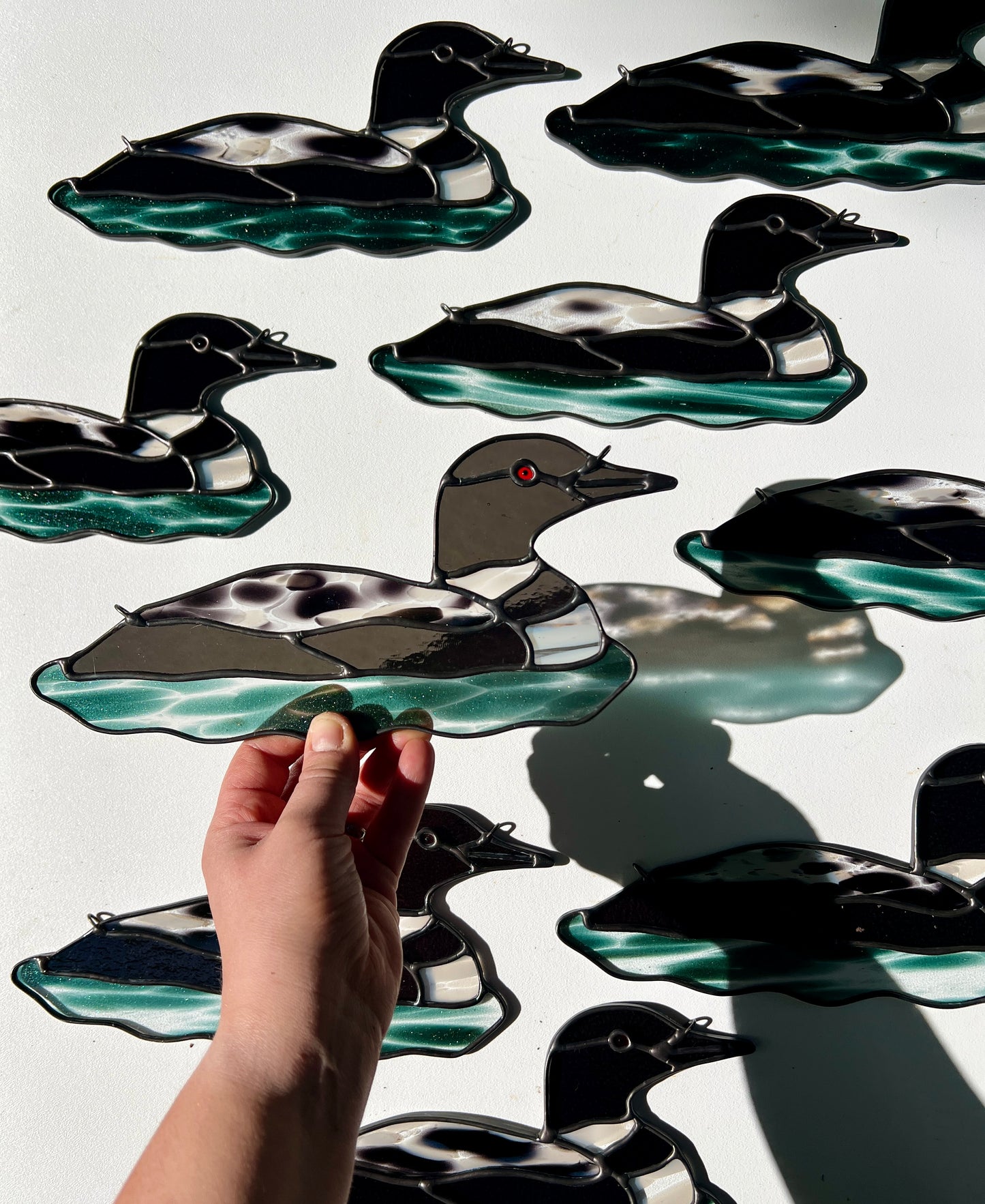 a hand holding a stained glass loon above a white surface with several other similar loon suncatchers arranged in the background