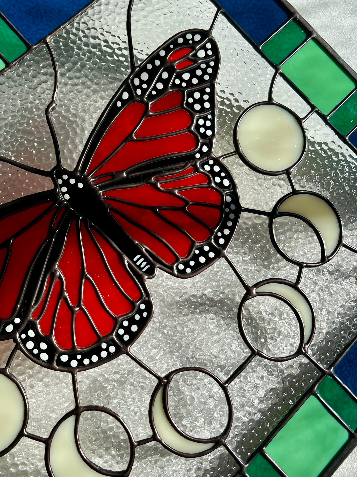 Close up of a large stained glass panel with a thick blue and green geometric border and a monarch butterfly and moon phases in the center, with textured clear glass in the background.
