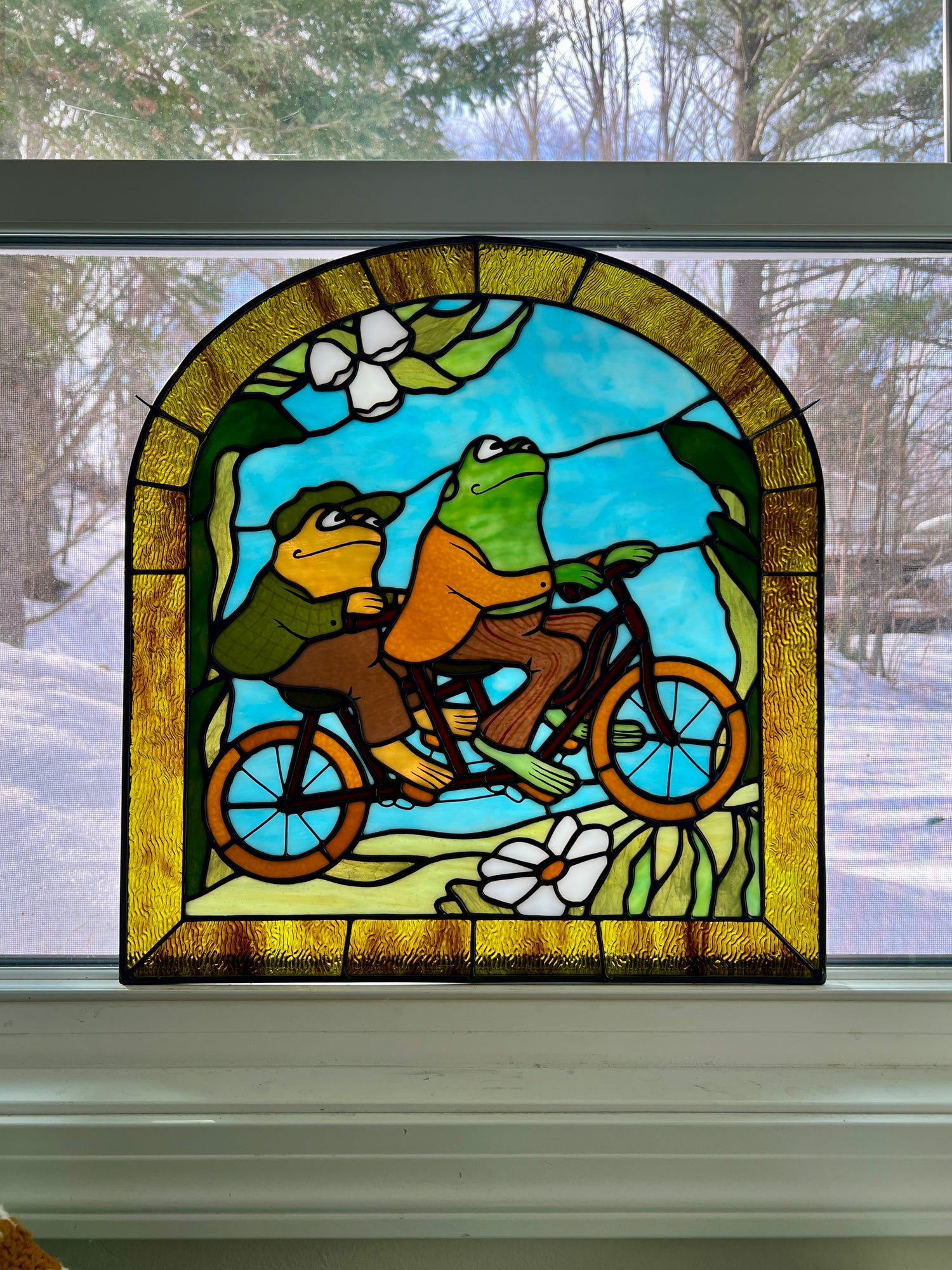 stained glass panel depicting frog and toad riding a bicycle resting on a window sill