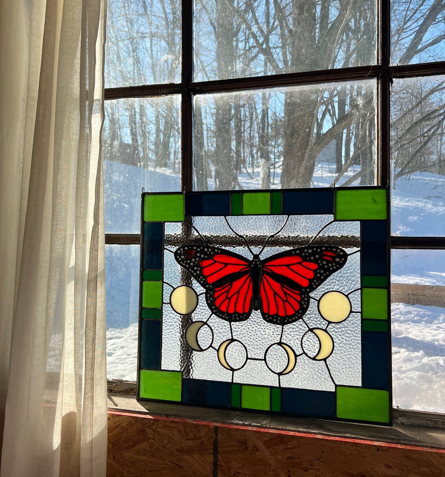 A large stained glass panel with a thick blue and green geometric border and a monarch butterfly and moon phases in the center, with textured clear glass in the background. The panel is resting in a window with bright sunlight providing backlighting. There are bare trees and snow on the ground in the background.