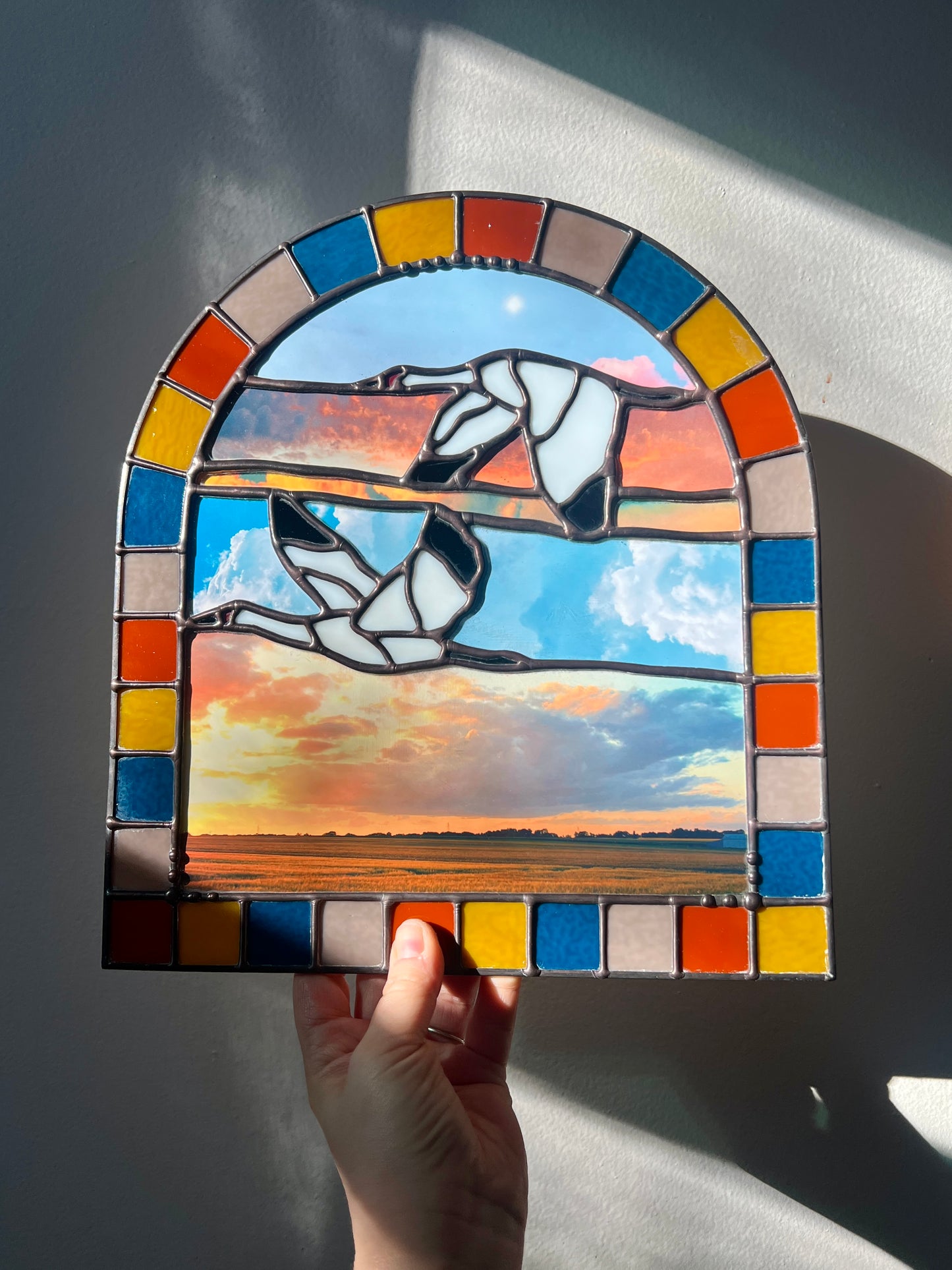 Stained glass panel with a landscape design created by collaged photographs and two cranes flying held by a hand against a neutral background
