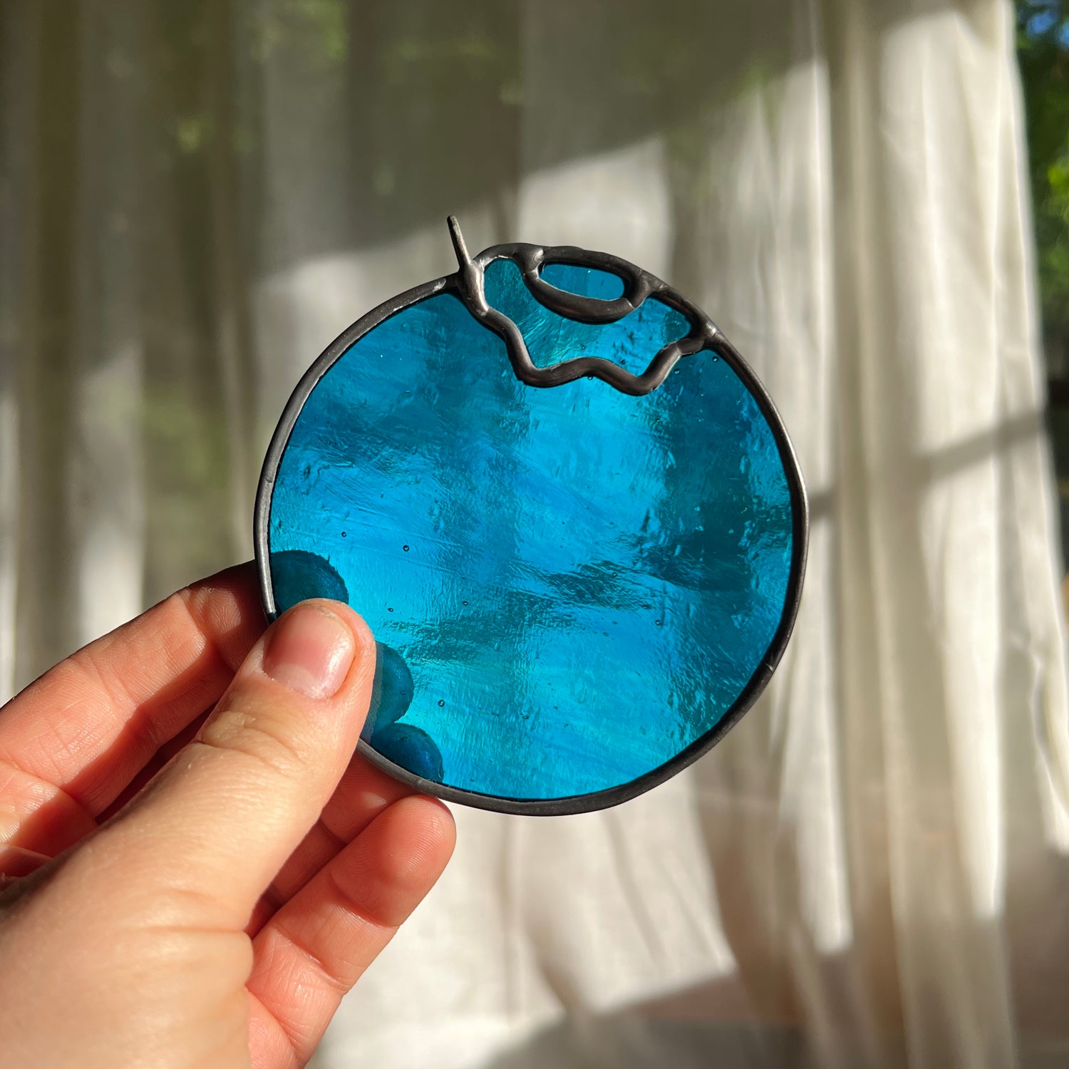 hand holding a wispy blue stained glass blueberry in front of a window