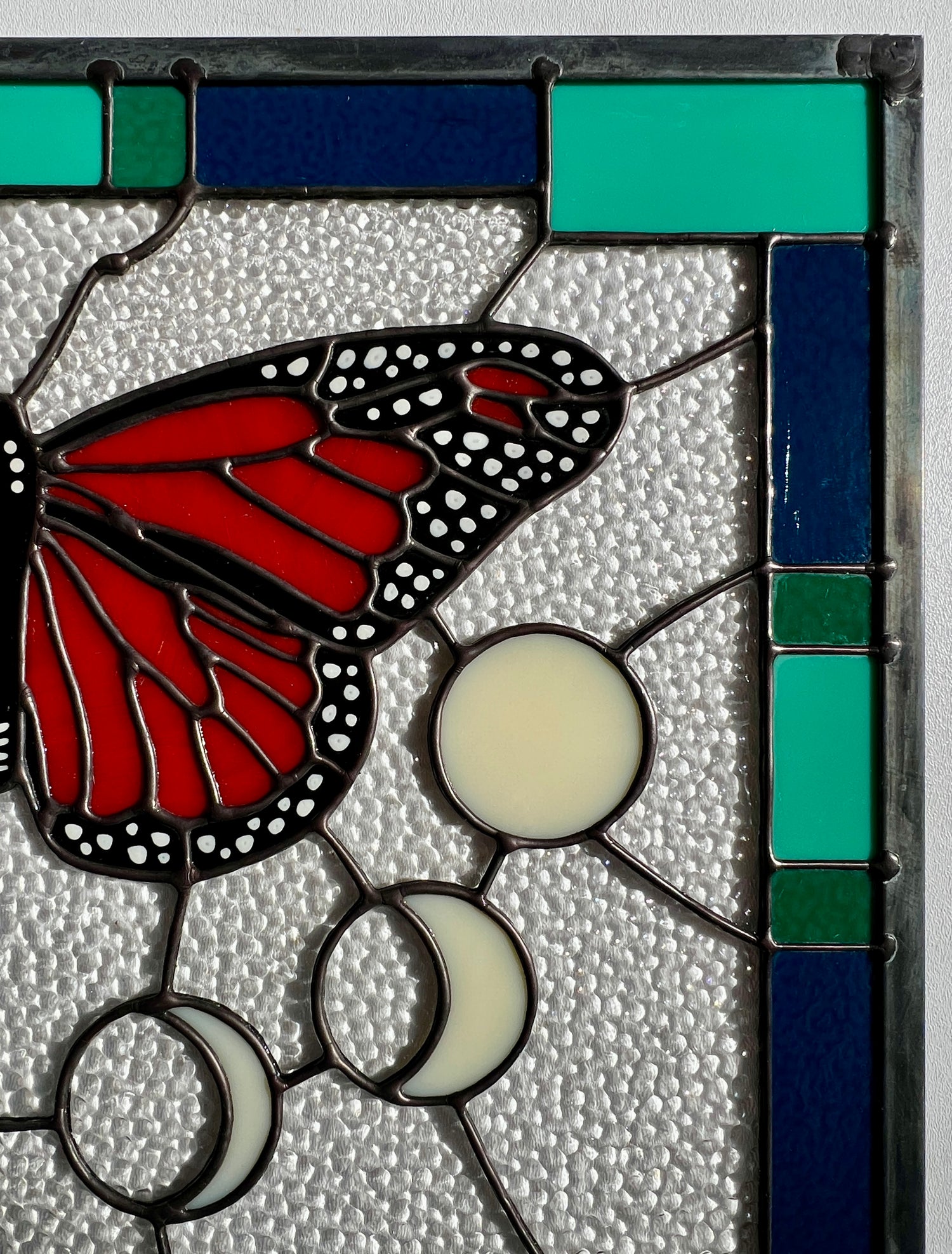 Close up of the upper right hand corner of a rectangular stained glass panel with a thick blue and green geometric border and a monarch butterfly and moon phases in the center, with textured clear glass in the background.