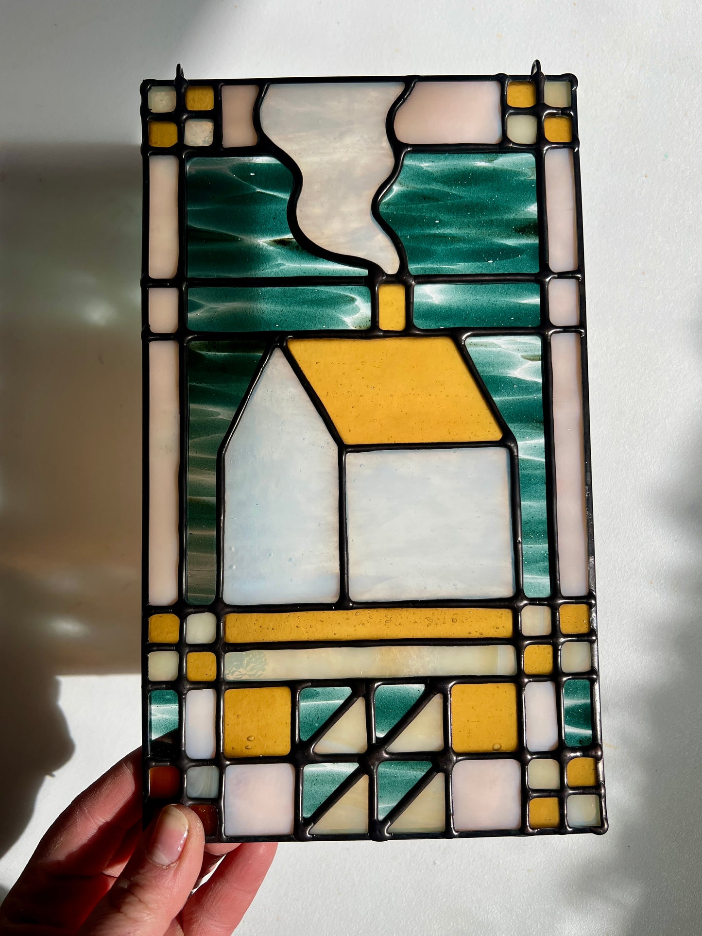 Stained glass textile inspired panel depicting a home in the center in shades of teal, amber, white, and beige, held by a hand in front of a white surface
