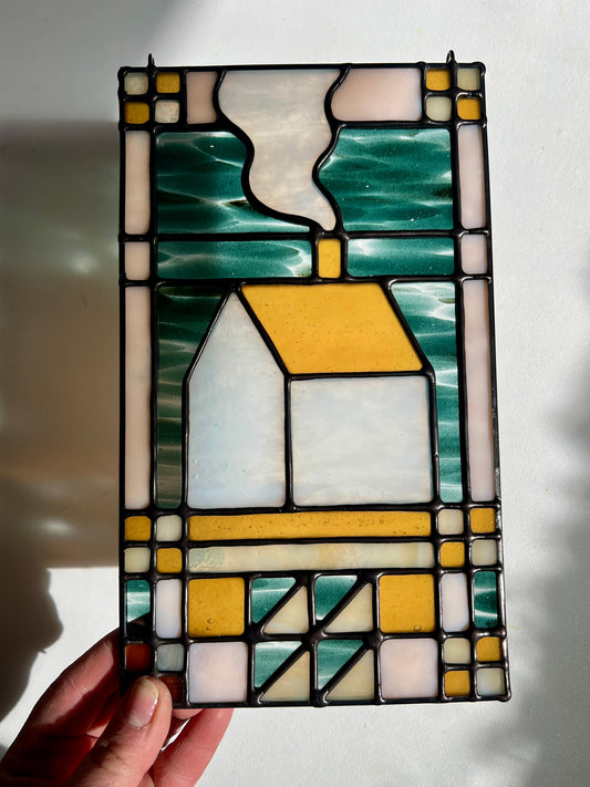Stained glass textile inspired panel depicting a home in the center in shades of teal, amber, white, and beige, held by a hand in front of a white surface