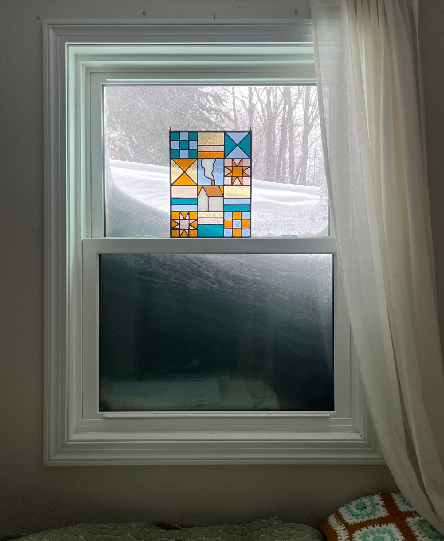 Stained glass textile inspired panel depicting a home in the center in shades of teal, pink, white, blue, and yellow,  resting in a window with an enormous snowbank outside.