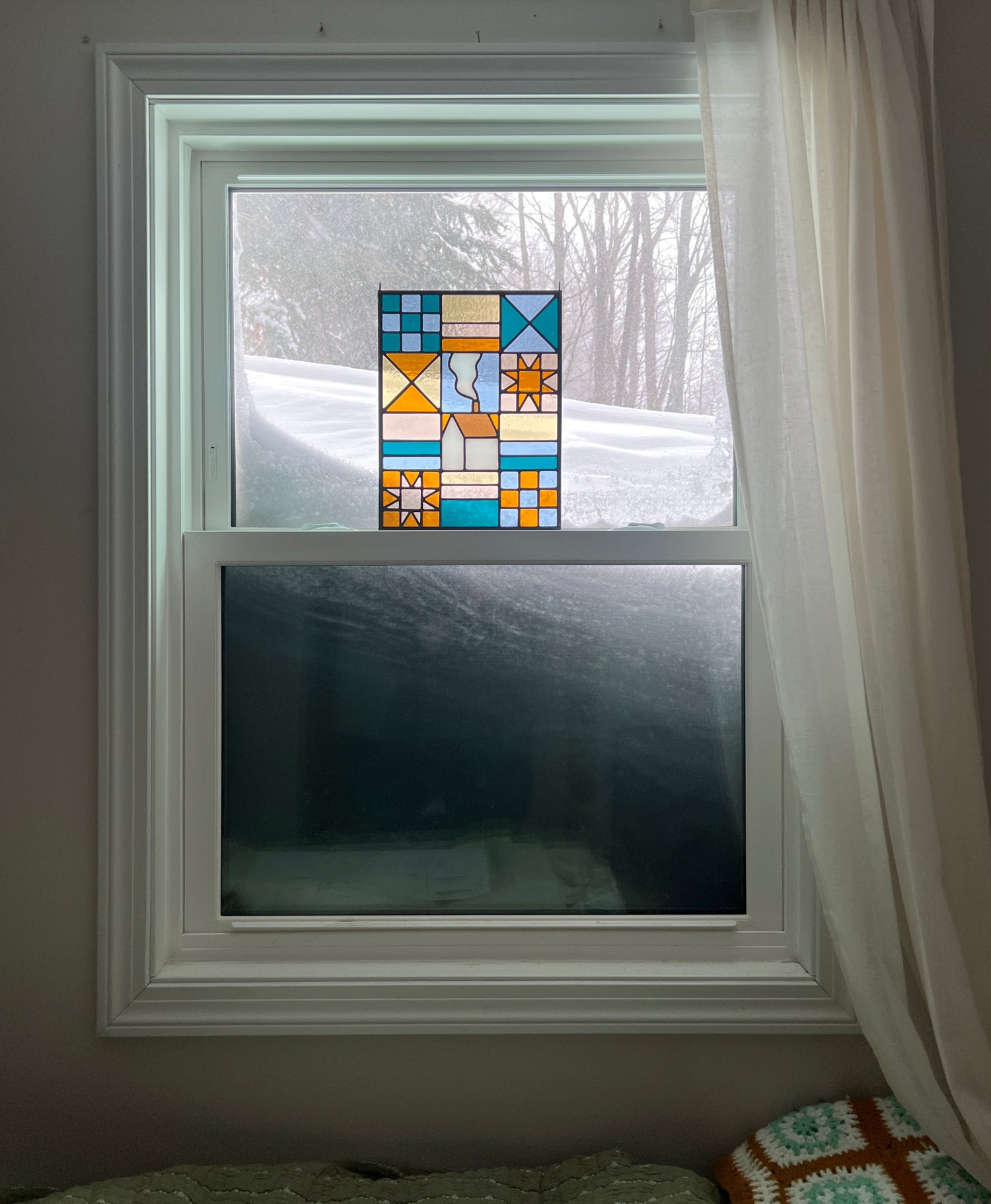 Stained glass textile inspired panel depicting a home in the center in shades of teal, pink, white, blue, and yellow,  resting in a window with an enormous snowbank outside.