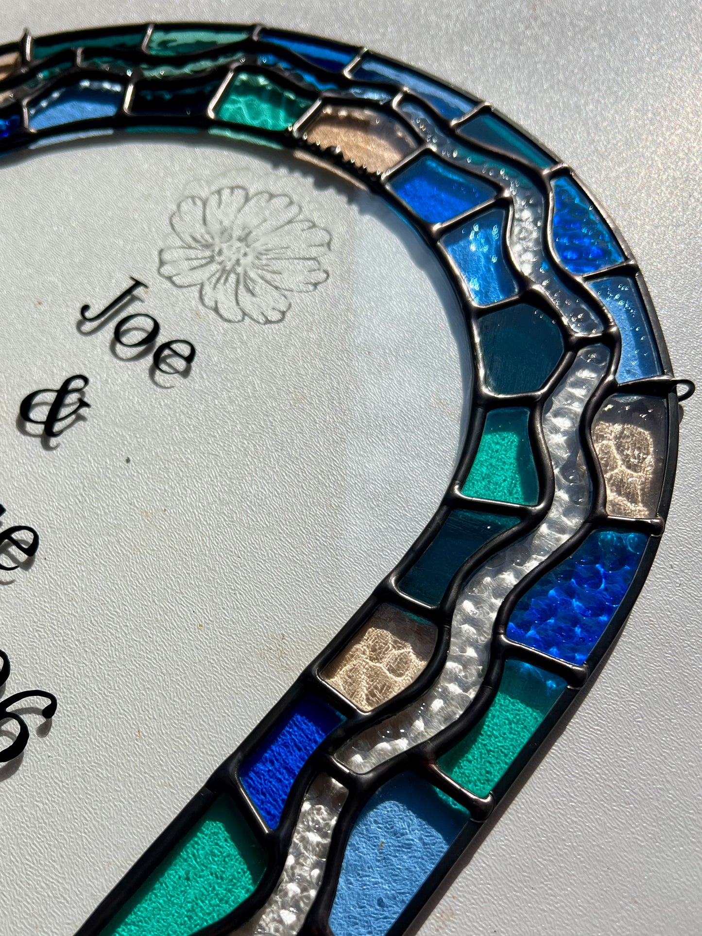 Close up of a stained glass panel in the shape of an arch with a multicolor blue, teal, turquoise, and pink border. Within the border is squiggle detail in clear textured glass. The center of the arch is clear uncolored glass, with personalized text and hand etched details of a cosmos flower. The photograph is taken at an angle against a white background to show smooth solder details. 