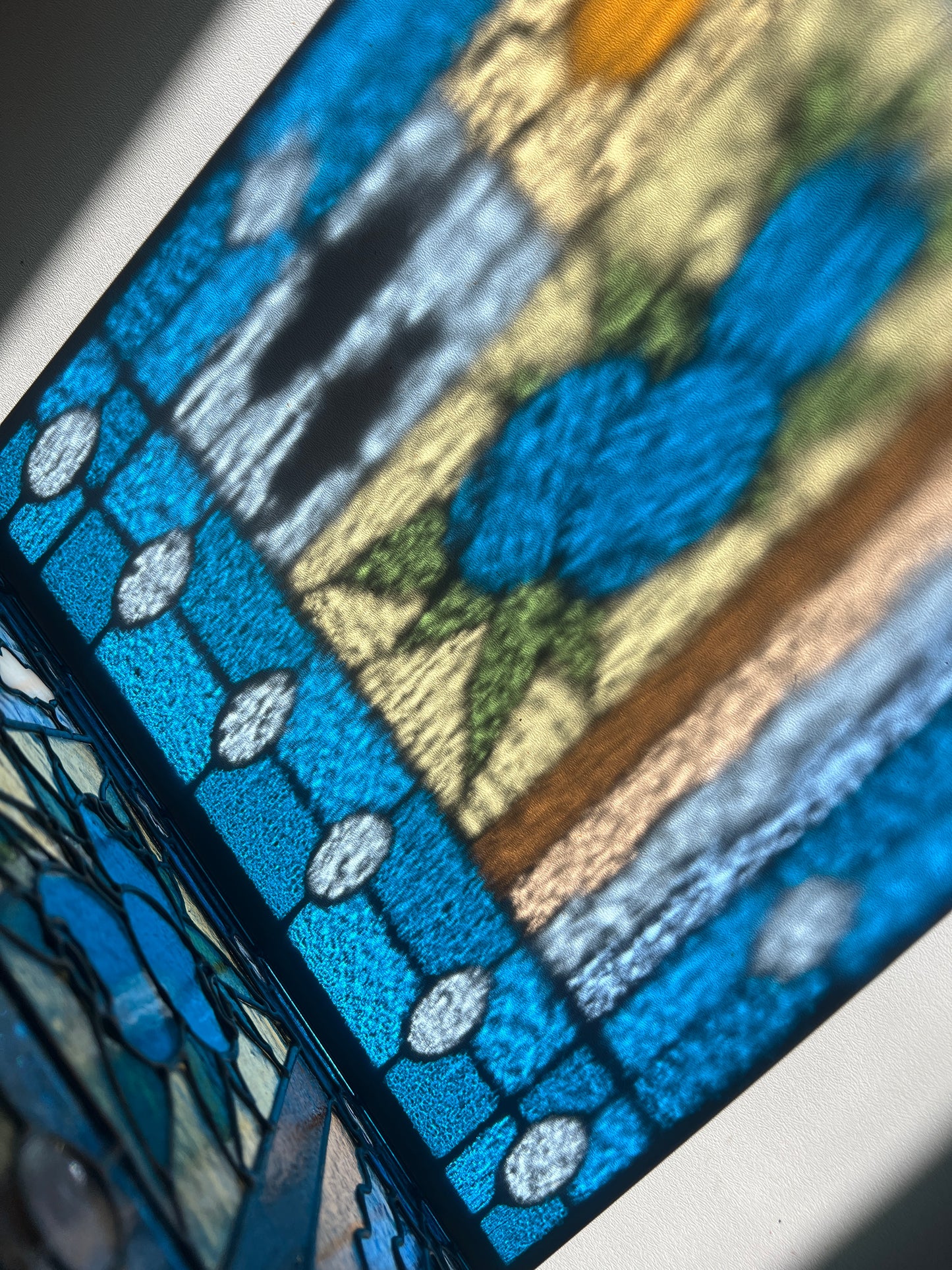 close up of a refraction of a stained glass panel with a simple compartment style design featuring clouds, sun, water, and a small bunch of blueberries in the middle with a blue border