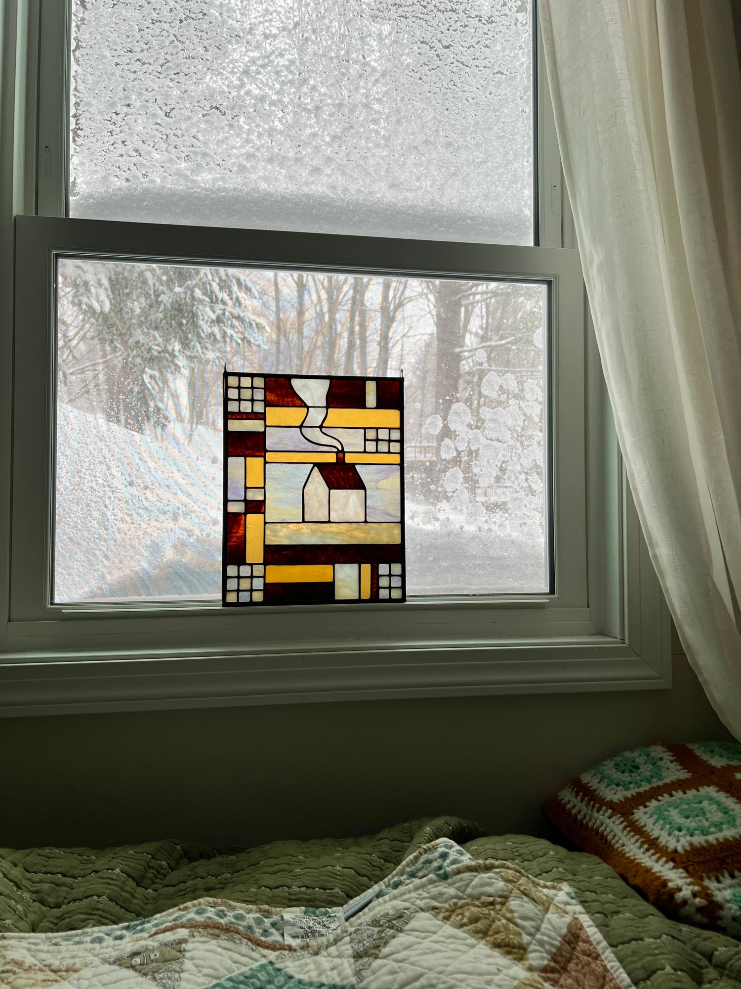 Stained glass textile inspired panel depicting a home in the center in shades of purple, white, brown, and amber, resting in a window with a snowy scene outside and cozy blankets in the foreground