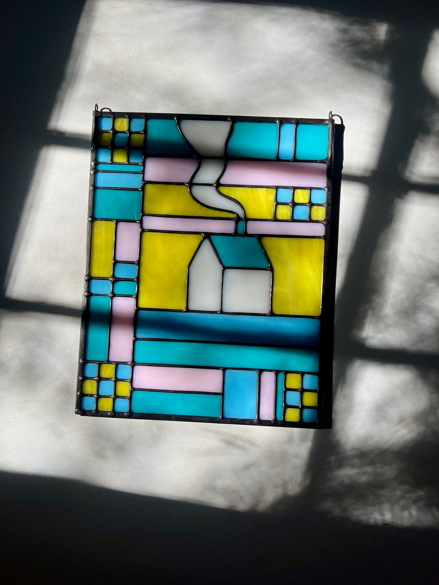 Stained glass textile inspired panel depicting a home in the center in shades of teal, pink, white, blue, and yellow, resting on a white surface with bright sunlight and shadows flowing across it.
