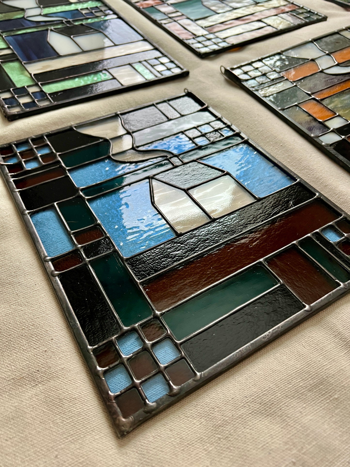 Stained glass textile inspired panel depicting a home in the center in shades of teal, brown, and blue, photographed at an angle to highlight smooth solder lines