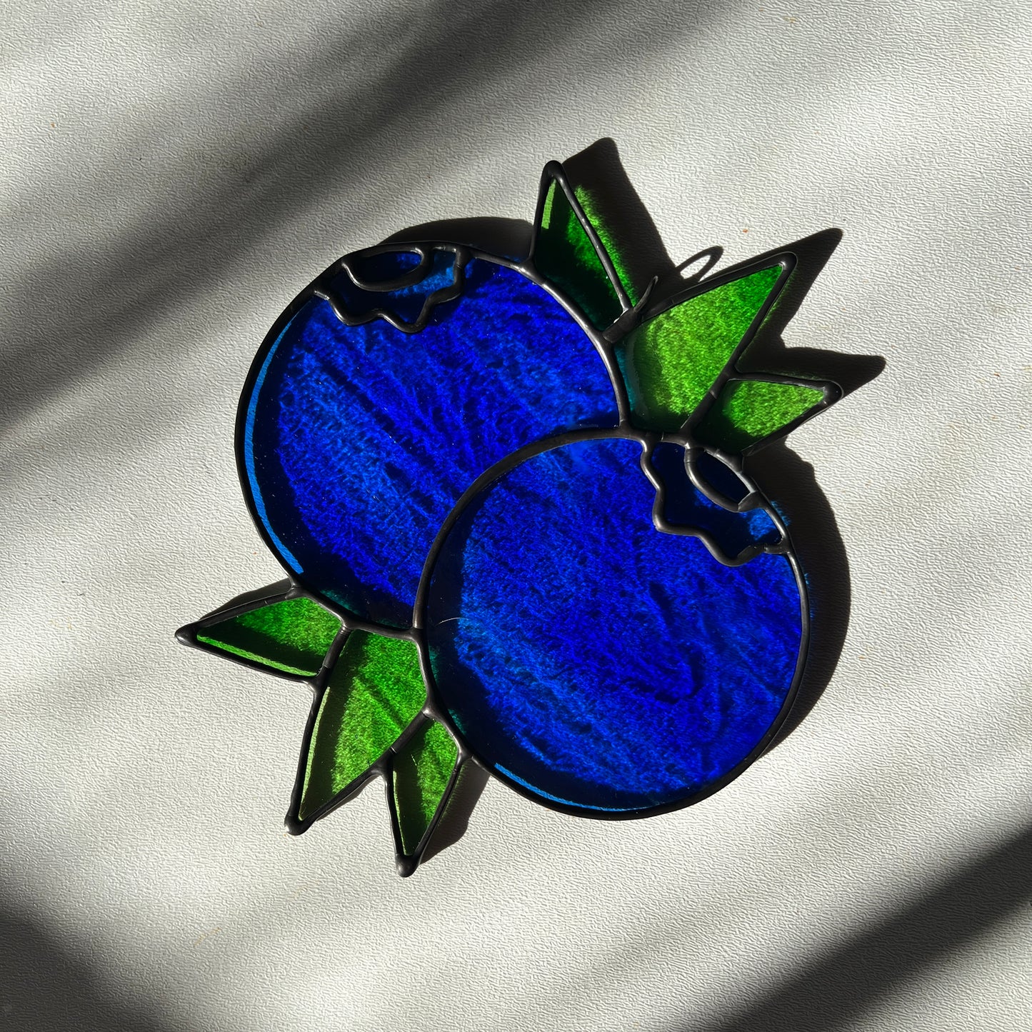a textured blue stained glass suncatcher depicting a small bunch of blueberries with leaves resting on a white surface