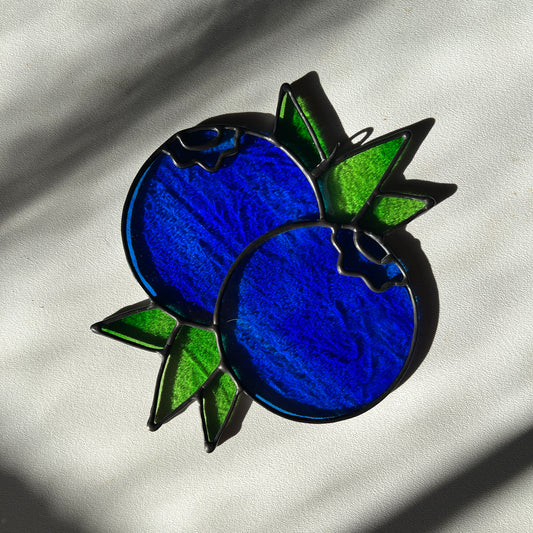 a textured blue stained glass suncatcher depicting a small bunch of blueberries with leaves resting on a white surface