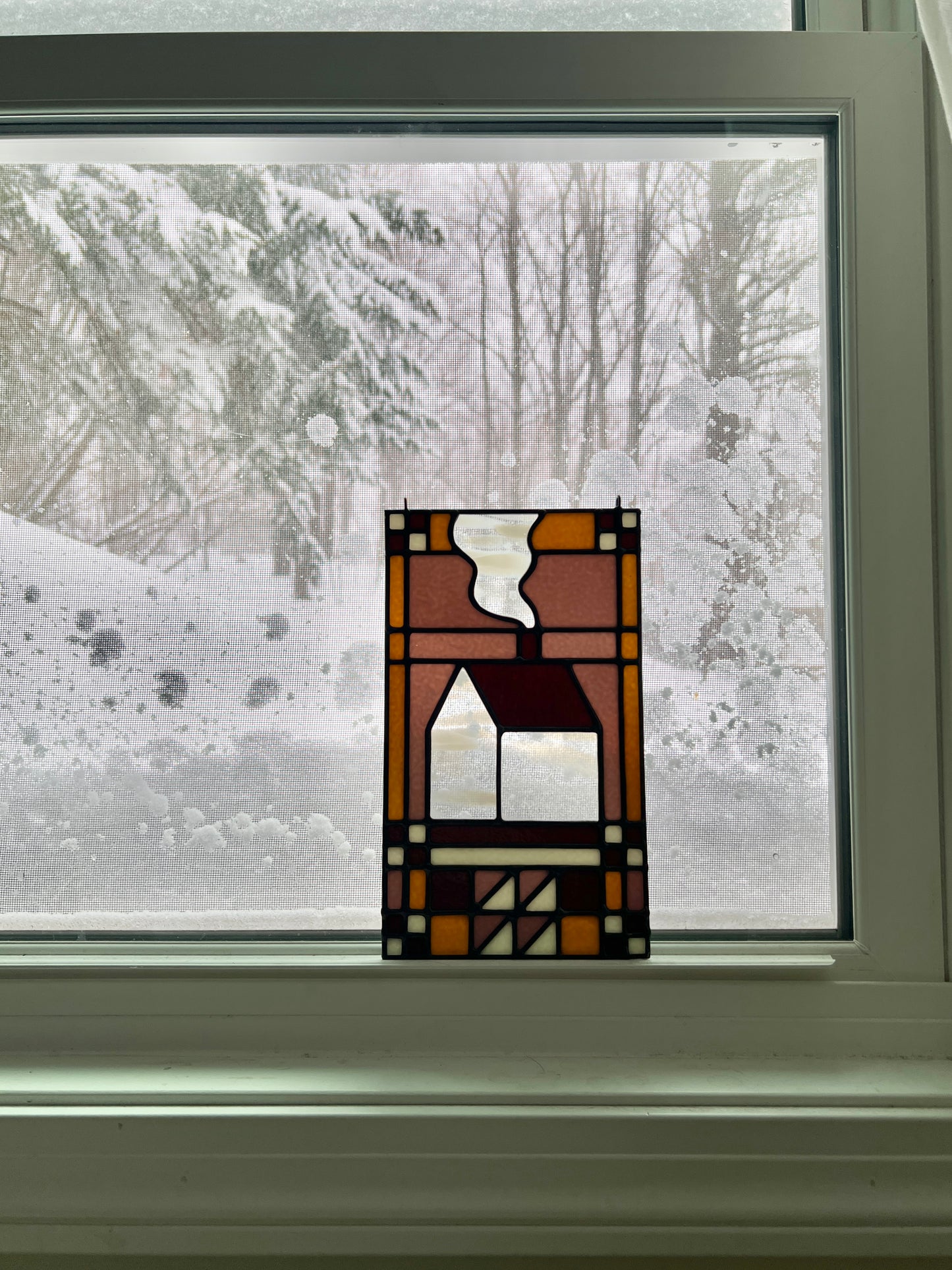 Stained glass textile inspired panel depicting a home in the center in shades of purple, white, brown, and bronze, resting in a window with a snowy scene outside