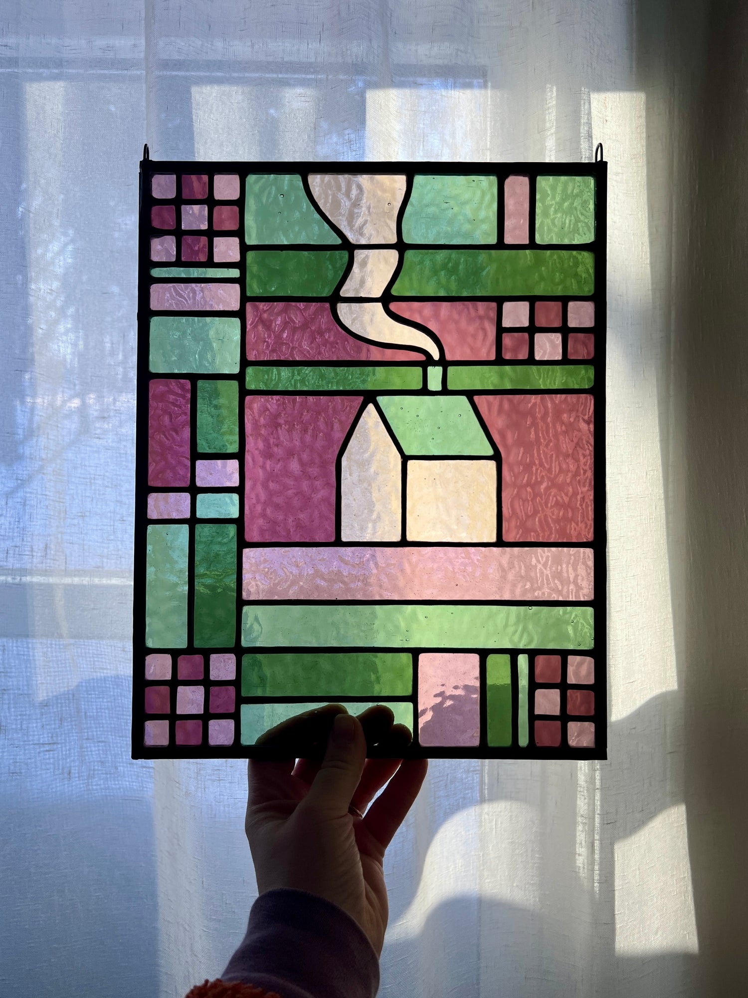 Stained glass panel with geometric textile inspired design with a cozy home depicted in the center, in shades of green and pink, held by a hand in front of softly lit sheer white curtain